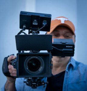 A man in an orange Texas Longhorns ball cap holding a professional video camera