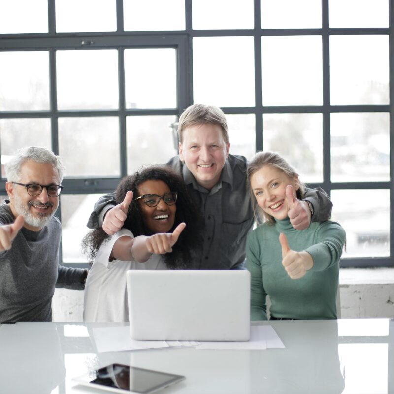 Two men and two women sitting in front of a laptop and posing with their thumbs up
