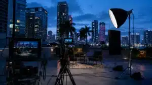 A professional B2B video shoot on a downtown Miami office rooftop at blue hour: a cinema camera on a tripod frames an empty interview chair, a softbox glows, and the illuminated Brickell skyline sits behind